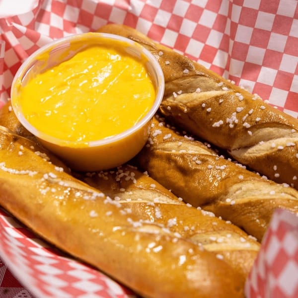 Salted pretzel sticks and a cup of bright yellow cheese dip in a red and white checkered basket.